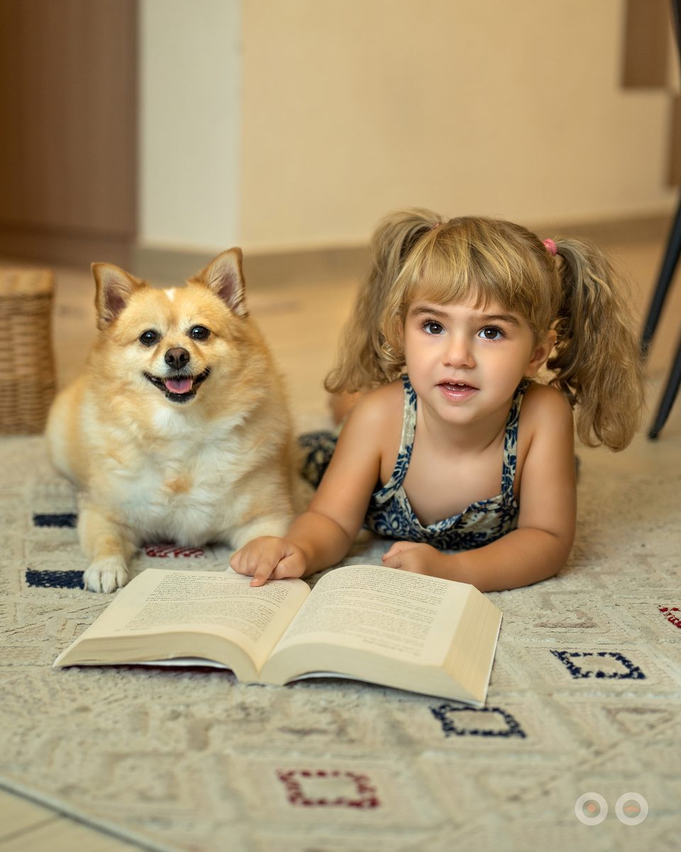 A small girl lying on the rug reading, a pomeranian settled beside her.